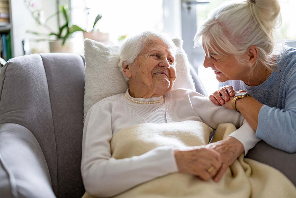 An elderly woman lies on a couch under a blanket while another older woman leans in, smiling and holding her hand in a well-lit room.