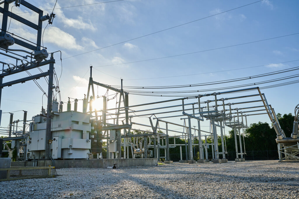 Outdoor electrical substation with transformers, circuit breakers, and power lines on a gravel lot, with the sun partially visible behind the equipment.