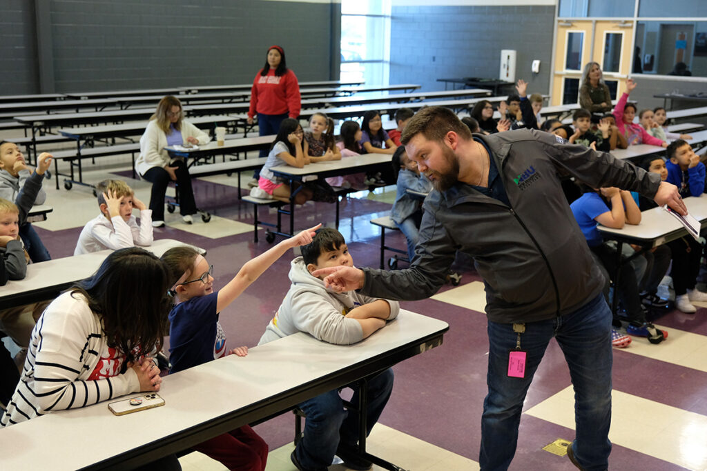 A man speaks with students seated at tables in a cafeteria while several children raise their hands to participate.