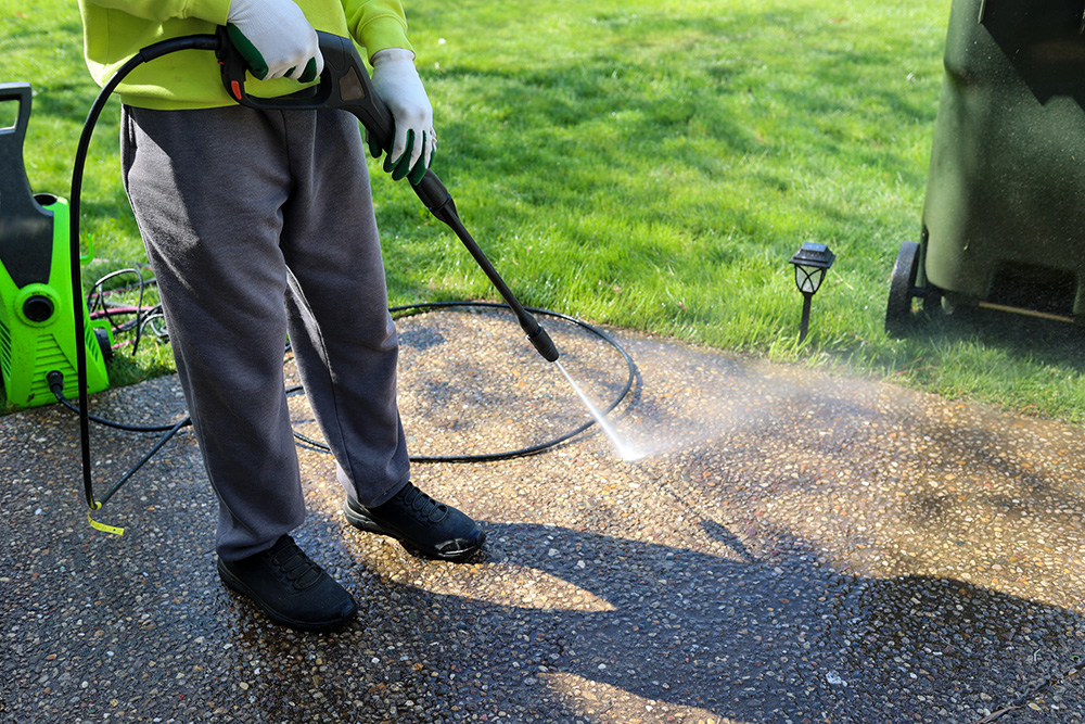 Person pressure washing a concrete patio outdoors on a sunny day, wearing gloves, dark shoes, and gray pants. Grass and cleaning equipment are visible in the background.