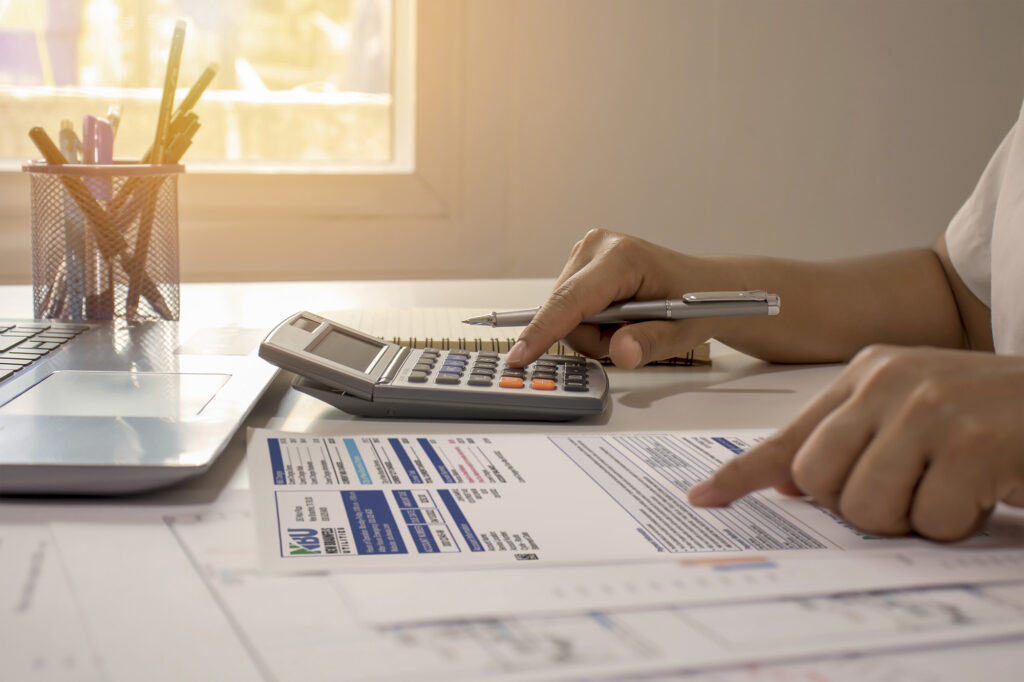 Person using a calculator and reviewing a printed financial document at a desk with a laptop and pen holder nearby.