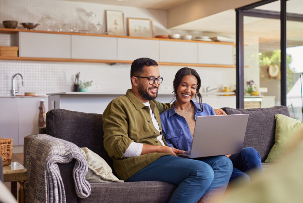 A man and woman sit together on a couch, smiling while looking at a laptop in a modern living room and kitchen setting.