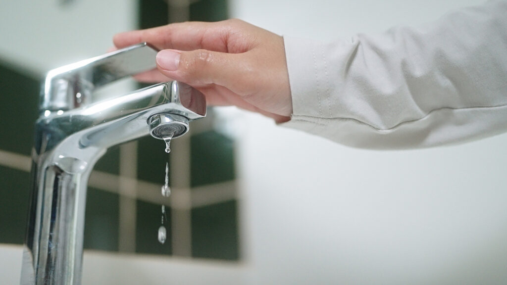 A hand turning off a modern silver faucet with water dripping from the spout.