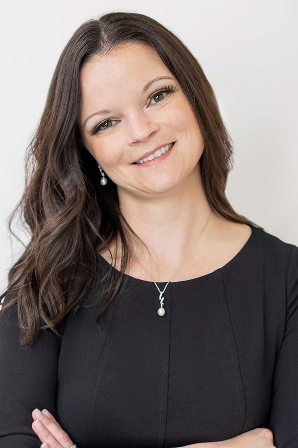 Woman with long brown hair wearing a black top, pearl necklace, and earrings, smiling with arms crossed against a plain light background.