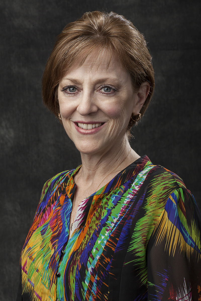 Portrait of a middle-aged woman with short brown hair, wearing a colorful patterned blouse, smiling, and posing in front of a dark background.