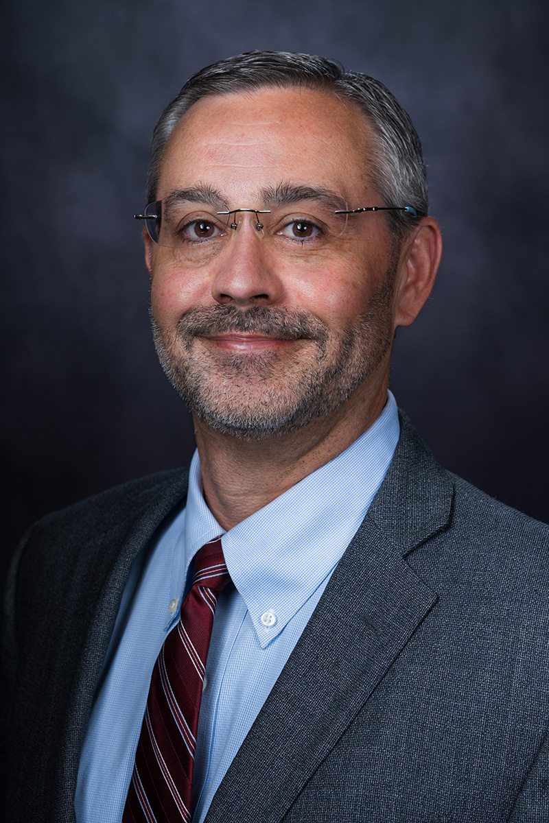 A middle-aged man with short gray hair, glasses, and a beard wears a gray suit, light blue shirt, and red striped tie against a dark background.