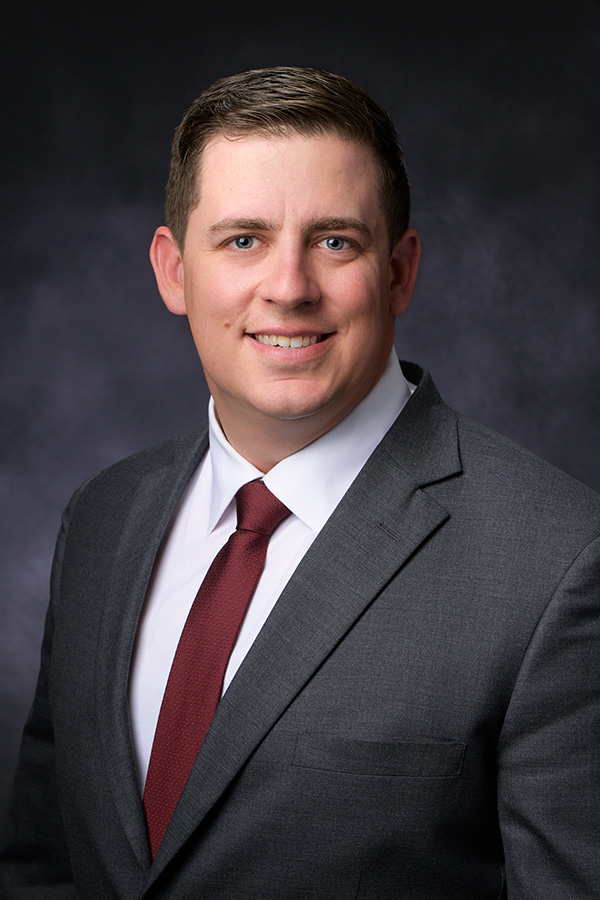 Man in a gray suit, white shirt, and red tie posing for a professional headshot against a dark, neutral background.