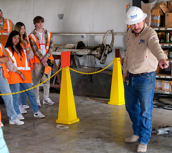 A man in a hard hat speaks to a group of students in safety vests inside a workshop, with tools and equipment visible in the background.
