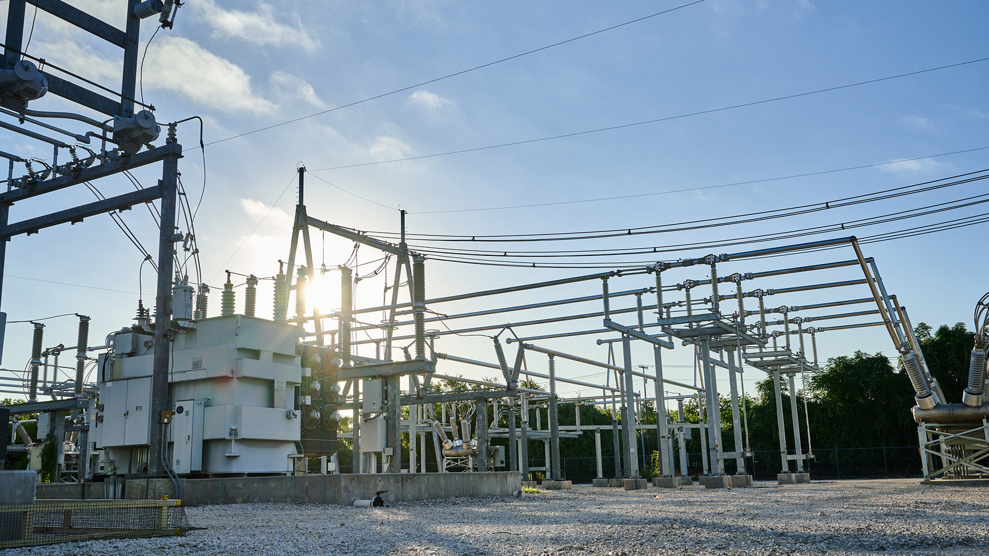 Outdoor electrical substation with transformers, circuit breakers, and power lines on a gravel lot, with the sun partially visible behind the equipment.