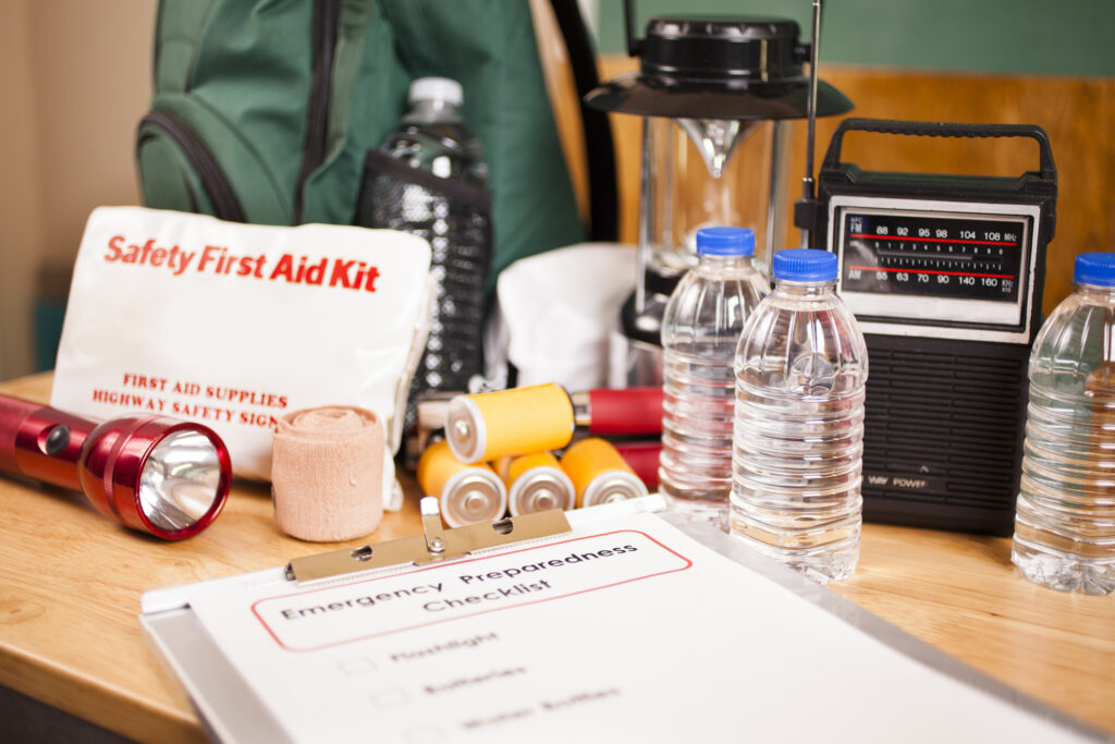Emergency preparedness checklist on clipboard and a large pile of supplies to be used in case of a natural disaster (hurricane, flood, earthquake, etc.) including: flashlight, batteries, water bottles, first aid kit, lantern, radio, backpack, mask.  No people.
